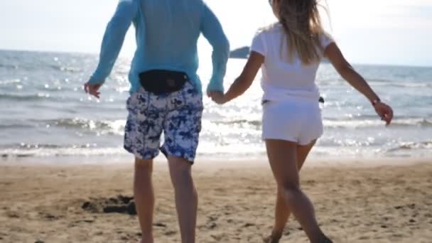 这对年轻夫妇手牵手 沿着海滩跑向大海 无法辨认的空气为自由和他们在海滨的暑假感到高兴 游客们在海边享受着新的风景 后视镜慢动作 — 图库视频影像