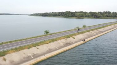 Motosikletlinin göl kenarındaki yolda motosikletiyle yarışırken çekilmiş bir fotoğrafı. Adam yaz günü baraj yolunda bisiklet sürüyor. Modern spor motosikletle nehir köprüsünün üzerinden uçuyor. Üst görünüm.