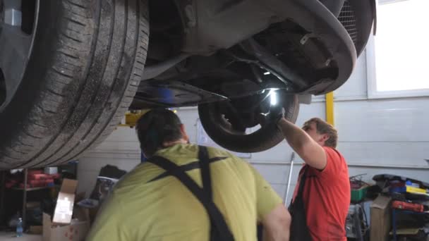 两名机械师检查汽车停放情况 车间里在起重车下面工作的专业修理工 男人们在讨论修理问题 汽车维修的概念 — 图库视频影像