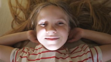 Yerde yatan ve odada kameraya bakan komik çocuk. Mutlu gülümseyen sarışın kız evde dinleniyor. Yüzündeki tatlı çocuğun olumlu duygularını kapat. Üst Görünüm Yavaş Çekim.