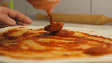Aşçının erkek kolu pizza hamuruna ketçap sürüyor mutfak masasında kaşık kullanıyor. Aşçının eli, mutfaktaki ahşap bir yüzeye hamur işine domates sosu sürüyor. Yemek hazırlama kavramı.