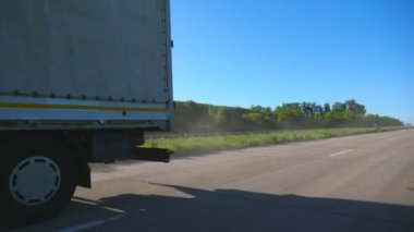 Kargo karavanı olan beyaz bir kamyon, güneşli bir günde otobanda araba sürüyor ve mal taşıyor. Kamyon arka planda güzel bir manzarayla kırsal alanda ilerliyor. Yavaş çekim ön görünümü Kapat.