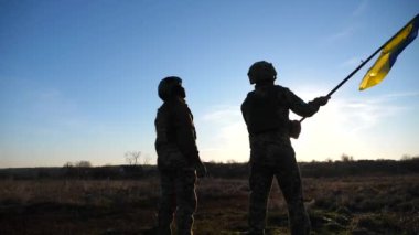 Ukrainian army soldiers stands outdoor at sunset time and waves flag of Ukraine. People in military uniform lifted up flag against blue sky. Victory at war. Resistance to russian invasion. Slow motion