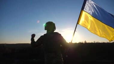 Soldier of ukrainian army stands at hill and holds waving flag of Ukraine. Man in military uniform and helmet lifted up flag against sunset. Victory against russian aggression. Invasion resistance.