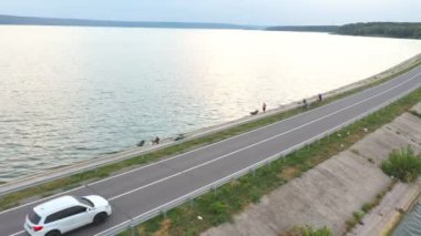 Beyaz arabanın havadan çekilmiş görüntüleri yaz günü baraj yolundan geçer. SUV göl ya da göl kenarındaki yolda gidiyor. Oto üzerinden uçmak nehir köprüsünden geçer. Hafta sonu seyahati kavramı. Üst görünüm