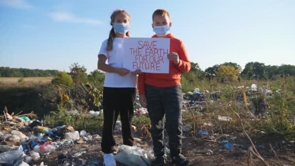 小男孩和小女孩手握环保运动的海报 拯救地球 戴着防护面罩的小生态志愿者看着摄像机 站在垃圾堆的后面 靠近点 — 图库视频影像