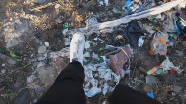 Spor ayakkabılı POV erkek ayağı çöp sahasında çöp kutusuna basıyor. Kırsal kesimdeki hurdalıkta yürüyen genç adam bacakları. Çöp doğada serbestçe ulaşılabilir yerlere atılır. Çevre kirliliği sorunu