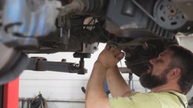 Tamirci araba tamirhanesinde ya da garajda araba tamir ediyor. Atölyede kaldırma aracının altında çalışan profesyonel sakallı oto tamircisi. Düzenli otomobil bakımı kavramı. Yavaş çekim.