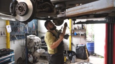 Otomobil tamircisi garajdaki bir kaldırma aracının altında çalışıyor. Profesyonel sakallı tamirci atölyede özel elektrikli aletle araba tamir ediyor. Otomobil bakımı kavramı. Yavaş çekim.