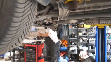 Garajda bir kaldırma aracıyla çalışan profesyonel bir oto tamircisi. Üniformalı sakallı tamirci atölyede elektrikli aletle araba tamir ediyor. Otomobil bakımı kavramı. Yavaş çekim.
