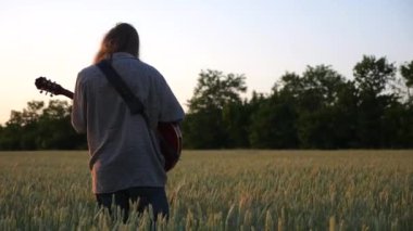 Erkek hipster buğday tarlasında elektro gitarla oynuyor. Uzun saçlı hippi adam arpa otlağında müzik aleti çalıyor. Genç adam doğada dinleniyor. Arka planda güzel bir gün batımı. Yavaş çekim