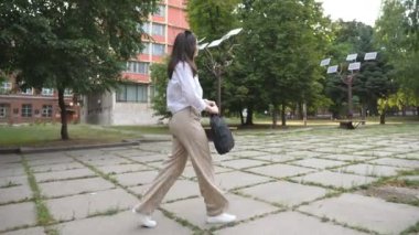 Resmi giyinmiş çekici iş kadınının şehir parkından geçerken görünüşü. Teneffüste çantalı genç bir kız dışarıda yürüyor. Başarı ve güven kavramı. Yavaş çekim.