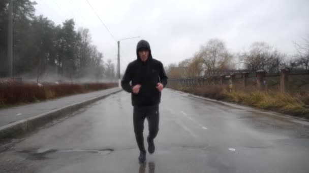 曇りの日にトレイルに沿ってジョギングするスポーツウェアの強いアスリート 雨の中アスファルト道路を走る若いスポーツマン 野外カーディオトレーニングを行っているスポーティな男性 健康的なアクティブライフスタイルのコンセプト — ストック動画