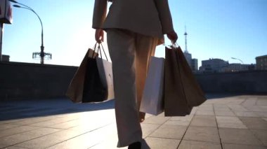 Young beautiful woman holds shopping bags walking at sunny city street after purchases. Fashionable girl carries paper packets going along urban square. Concept of leisure and free time. Slow motion.