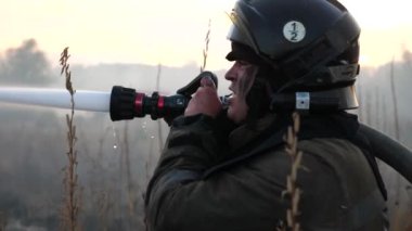 Üniformalı erkek itfaiyeci kırsal kesimdeki yangın hortumuyla yanan otları söndürüyor. Tam teçhizatlı profesyonel itfaiyeci çayırı ateşe veriyor. Kahramanlık mesleği ve yangın güvenliği kavramı.