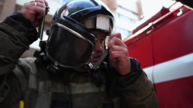 Male firefighter taking off helmet and balaclava against background of a fire engine. Young fireman removing equipment from himself after fire. Tired fireguard looking into camera near a big red truck