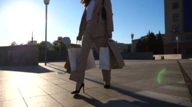 Elegant young lady holds shopping bags going at city square after purchases. Attractive woman carries paper packets walking along sunny urban street. Sales and discounts concept. Slow motion.
