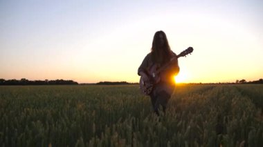 Uzun saçlı hippi adam yeşil arpa çayırında yürürken elektro gitarıyla çalıyor. Erkek hipster buğday tarlasında müzik enstrümanı çalıyor. Arka planda güzel bir gün batımı. Yavaş çekim.