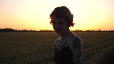 Buğday tarlasında dolaşan ve özgürlüğün tadını çıkaran dövmeli güzel punk kızı. Mutlu bayan hippi gün batımında yeşil arpa tarlasında yürüyor. Arka planda güzel manzaralı kırsal alan. Kapat.