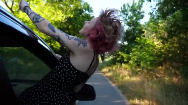 Otomobil camından sarkan pembe saçlı mutlu punk kızı kırsal kesimde kameraya bakıyor. Dövmeli kaygısız hippi kadın kır yolunda araba sürerken gezintiden zevk alıyor. Seyahat konsepti. Kapat.