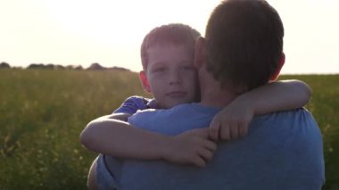 Küçük oğlunun elini tutan ve yeşil çayırlarda yürüyen genç bir baba. Küçük çocuk kameraya bakıyor ve gün batımının arka planında babasına sarılıyor. Aile dinlenmesi kavramı. Kapat..