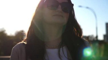 Portrait of beautiful lady in sunglasses walking at sunny day. Attractive brunette girl looks into camera going along city street. Businesswoman commuting to work. Concept of urban lifestyle. Close up