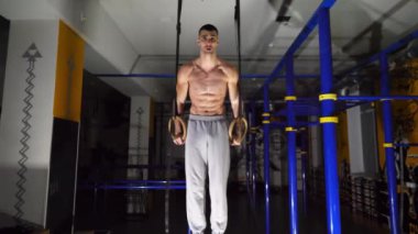 Modern spor salonunda kaslı üstsüz sporcu jimnastik yüzükleriyle şınav çekiyor. Güçlü atlet içeride sıkı egzersiz yapıyor. Spor kulübünde genç erkek jimnastikçi eğitimi. Sağlıklı ve aktif yaşam tarzı kavramı.