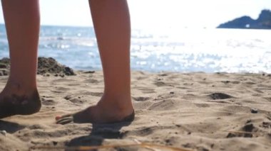 Sahilde altın kumda yürüyen kadın ayakları ve arka planda deniz manzarası. Deniz kıyısında yürüyen genç kadın bacakları. Çıplak ayaklı kız okyanus kıyısında geziniyor. Yaz tatili kavramı.