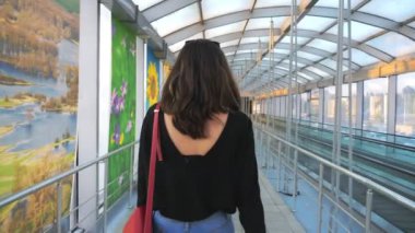 Yüksek topuklu ayakkabı giyen tanınmayan genç bir kız, cam terminal koridorunda bavuluyla yürüyor. Çekici, zayıf kadınların bavullarını tekerleklerle çekmesi. Bayan tökezleyecek. Yavaş çekim.