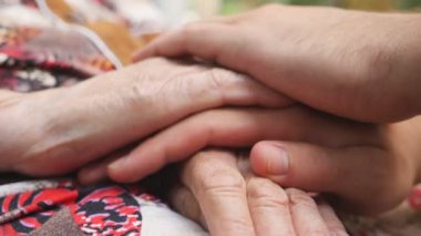 Genç erkek ve kırışık ellerin yakınlaşması ve dışarıda birbirini okşaması. Torun ve büyükanne dışarıda vakit geçiriyorlar. İlgi ve sevgi kavramı. Yan görünüm Yavaş çekim.