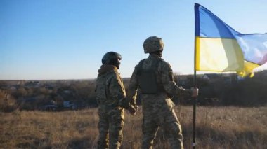 Kamuflaj üniformalı genç askerler gün batımına karşı ellerinde Ukrayna bayrağıyla çayırda duruyorlar. Ukrayna ordusundan bir çift, savaş alanında el ele tutuşan bayrağı kaldırdı. Dolly vurdu..