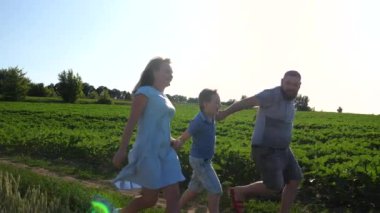 Mutlu aile çayırlarda koşuşturuyor ve el ele tutuşuyorlar. Anne ve baba ve küçük oğlu dışarıda eğleniyorlar. Tasasız ebeveynler ve küçük çocuk güneşli yaz gününde birlikte vakit geçiriyorlar. Yavaş çekim.