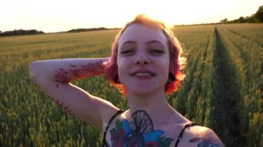 Pembe saçlı, kameraya bakıp Arpa Çayırı 'ndaki video için video kaydeden neşeli hippi kız. Kulak tünelleri olan mutlu punk kızı gün batımında kendini akıllı telefonuyla buğday tarlasında filme çekiyor..
