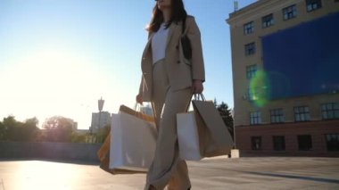 Güzel takım elbiseli genç bayan alışveriş sonrası kaldırımda yürüyen alışveriş çantaları tutuyor. Zarif bir kadın şehir caddesinde kağıt paketler taşıyor. Satış ve indirim kavramı. Düşük görünüm Yavaş çekim.