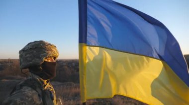 Ukrayna ordusunun askeri gün batımında mavi-sarı bayrak sallayarak tepenin zirvesinde duruyor. Kamuflaj üniforması ve Ukrayna bayrağı taşıyan genç bir erkek asker. İstila direnişi kavramı.