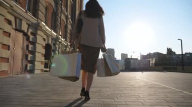 Şık bir kadının arka görünüşü, şehir caddesinde yürüyen kağıt paketler taşıyor. Zarif genç kız alışveriş sonrası kaldırıma çıkan alışveriş torbalarını tutuyor. Mevsimlik satış ve indirim kavramı. Yavaş çekim.