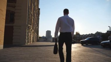 Şehir caddesinde evrak çantalı başarılı bir iş adamının arka görüntüsü. Resmi kıyafetli genç girişimci kaldırımda işe gidiyor. Kendine güvenen bir erkek yönetici işe gidip geliyor. Yavaş çekim.