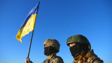 Ukrayna ordusunun genç askerleri ulusal bayrağıyla tepenin zirvesinde dikiliyorlar. Kamuflaj üniformalı kadın ve adam mavi gökyüzüne karşı Ukrayna bayrağını kaldırmış uzaklığa bakıyor. Kapat..