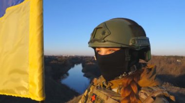 Ukrayna ordusunun kadın askeri doktoru bayrağı tutuyor ve gün batımını seyrediyor. Kamuflaj üniformalı ve kasklı genç bir kız Ukrayna bayrağını tepede dalgalandırdı. Rus istilasına karşı direniş.