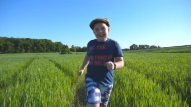 Mutlu çocuk Portresi güneşli sıcak bir günde yeşil buğday ile alanında çalışıyor. Yaz aylarında çayırda koşu ve eğlenen şapkalı gülümseyen çocuk. Çocukluk ve dinlenme kavramı. Yavaş çekim.
