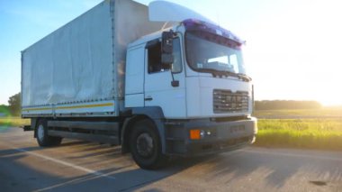 Kargo karavanı taşıyan bir kamyon otobanda sürüp mal taşıyor. Beyaz kamyon gün batımında kırsala doğru gidiyor. Parlak günbatımı ışığı arka planda parlıyor. Yan Görünüm Yavaş çekim.