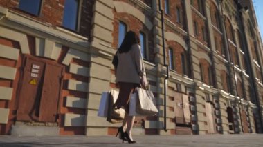 Modaya uygun bir kadın şehir caddesinde kağıt paketler taşıyor. Alışveriş sonrası kaldırımda yürüyen alışveriş torbaları tutan genç bir kız. Mevsimlik satış ve indirim kavramı. Dolly çekim ağır çekim.