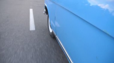 Point of view to front left tire of retro car. Wheel of old automobile riding on asphalt road. Blue vintage auto driving at the city street on summer day. Close up Slow motion.