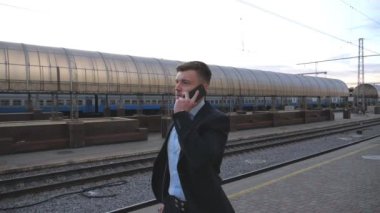 Successful businessman walking through railway station and talking on phone. Handsome man strolling near railway and having work conversation. Guy goes outdoor speaking on cellphone. Slow motion.