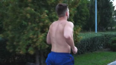 Kablosuz kulaklıklı genç kaslı sporcu yaz günü ormanda kaldırım kenarında koşuyor. Sportif yakışıklı adam doğada koşuyor. Sağlıklı aktif yaşam tarzı kavramı. Dolly vurdu..