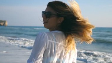 Güneşli bir günde sahilde yürüyen ve dönen genç bir kadın. Güneş gözlüklü güzel kız sarı saçlarına dokunuyor ve yaz tatilinin tadını çıkarıyor. Yaz tatili ya da tatil kavramı. Yaklaş. Ağır çekim.