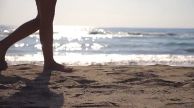 Kadın ayakları arka plan dalgalar ile güneşli bir günde deniz Sahilde yürüyüş. Genç kadın kum adım bacaklar. Yaz tatil veya tatil kavramı. Yan görünümü ağır çekim yakın çekim.