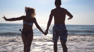 Genç mutlu çift sahilde koşuyor ve el ele tutuşuyorlar. Kız ve erkek yaz tatilinde ya da tatilde birlikte eğleniyorlar. Arka plan görünümünü kapat Yavaş çekim.