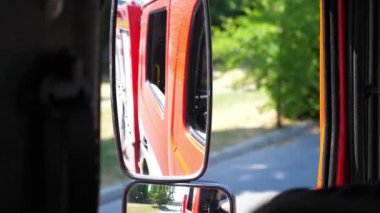 View to side mirror of fire engine riding to an emergency. Red big truck driving to place of a fire on sunny summer day. Concept of saving lives and heroic profession. Close up Slow motion.