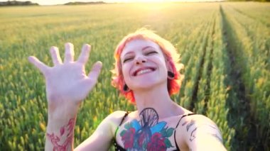 Pembe saçlı neşeli hippi kızın görüntülü konuşması akıllı telefondan ve buğday tarlasında el sallaması. Kulak tünelleri olan mutlu punk kadın kameraya bakıyor ve blog için video kaydediyor.